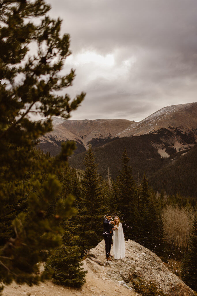 Couple popping champagne on a rocky overlook during a Winter Park elopement, surrounded by evergreen trees and Colorado mountain views.