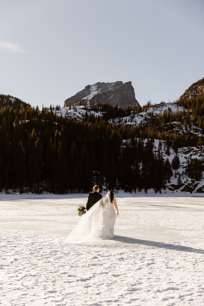 Bride and groom walking across a frozen lake during their December elopement in Rocky Mountain National Park with snow-covered mountains in the background.
