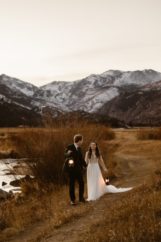 Bride and groom walking along a mountain trail at sunset during their December elopement in Rocky Mountain National Park holding lanterns.