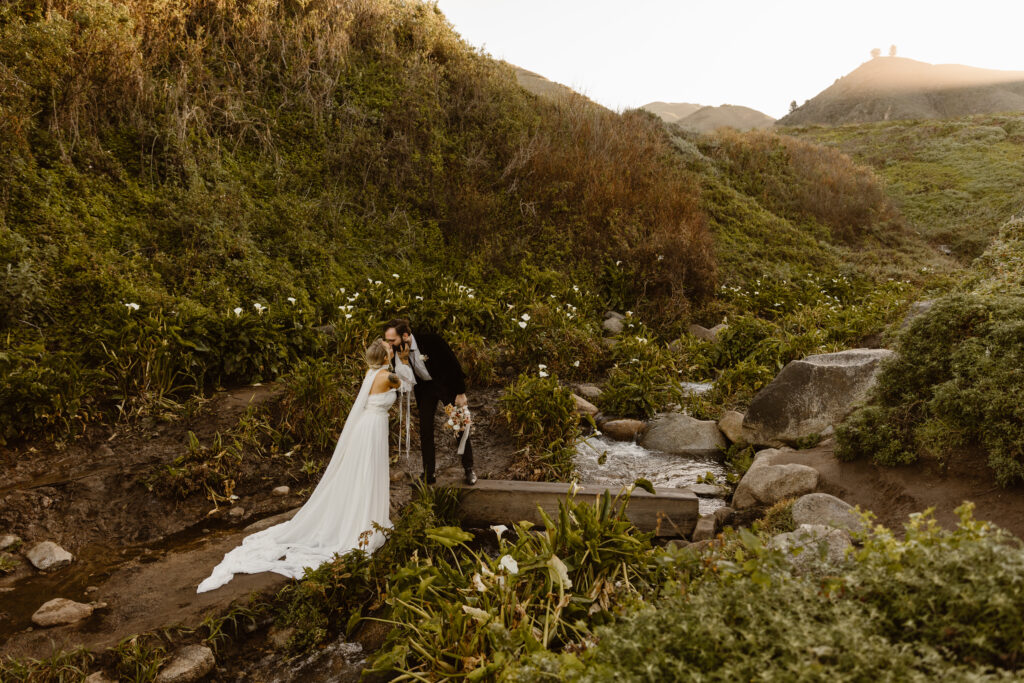 Bride leans in to kiss groom as he stands on a wooden plank over a creek surrounded by calla lilies in Big Sur