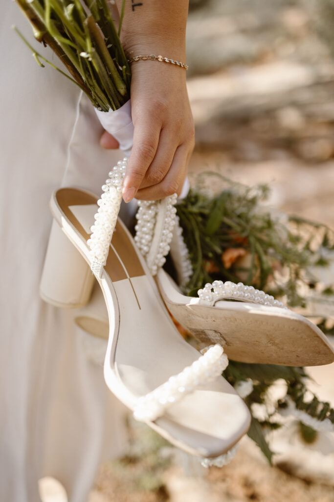 Bride holding pearl-strap heels and a bouquet during an elopement at Colorado National Monument, with soft desert tones in the background.