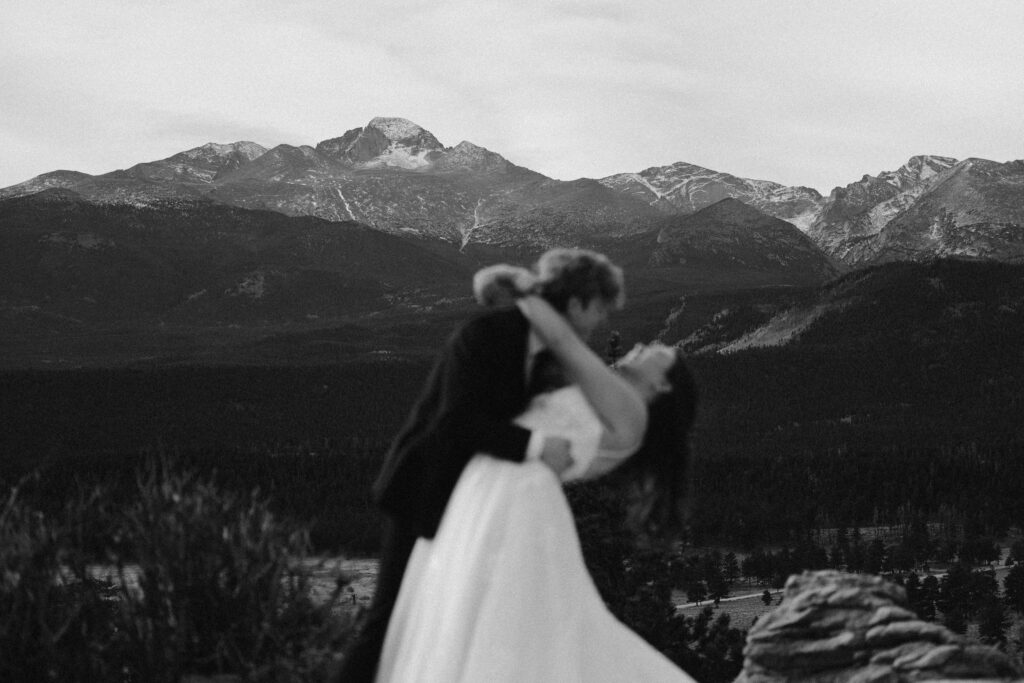Black and white blurred photo of a groom dipping the bride on a mountain overlook with Rocky Mountain National Park peaks in the background