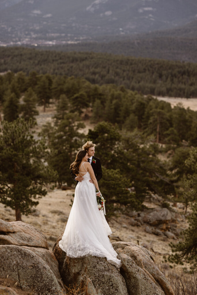 Bride and groom stand together on large rocks overlooking a forested valley in Rocky Mountain National Park during a November elopement