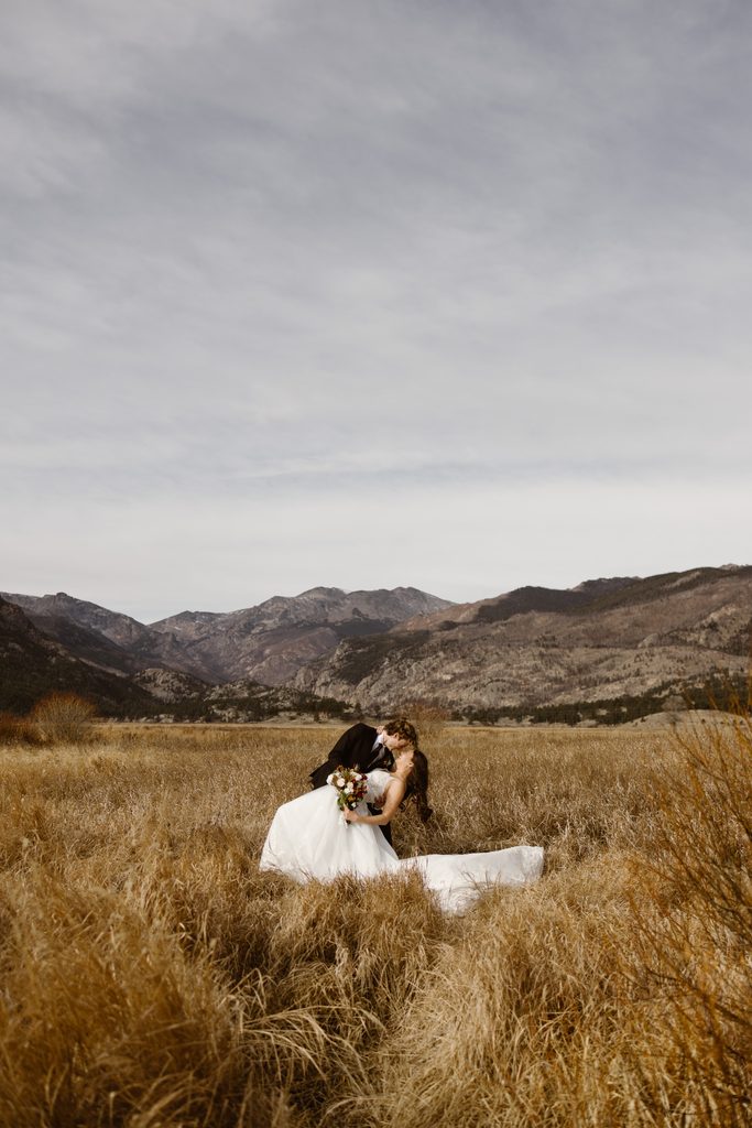 Wide view of groom dipping bride for a kiss in a golden meadow in Rocky Mountain National Park with expansive mountains behind them