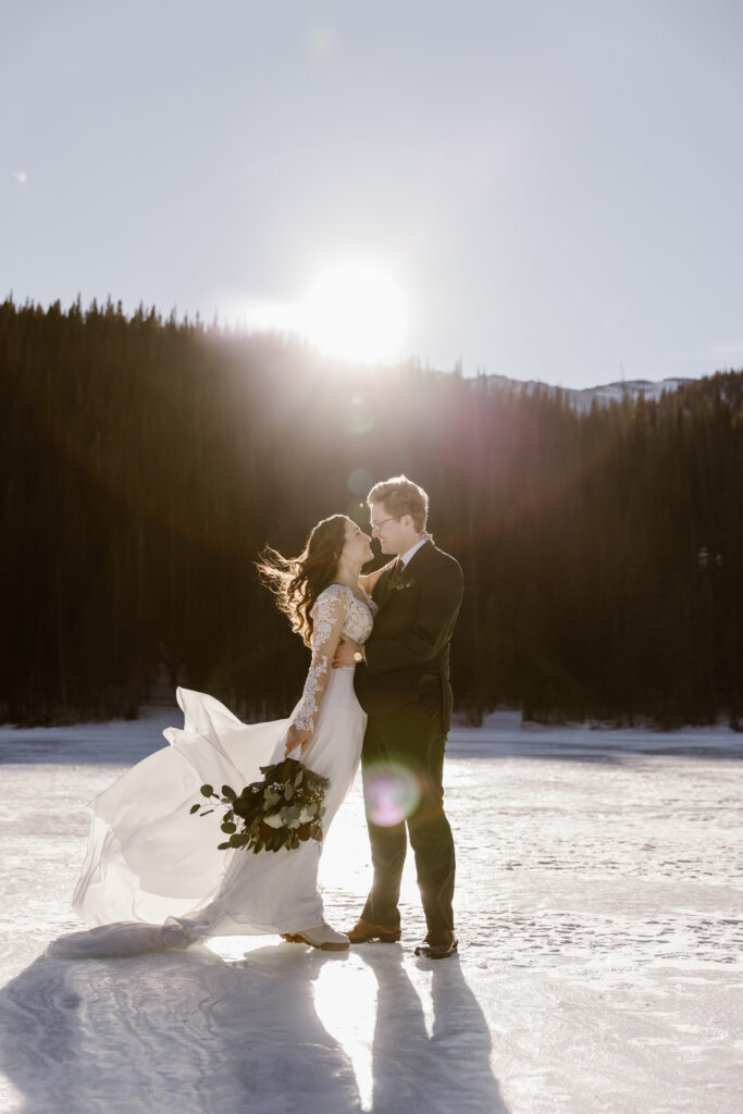 Bride and groom embrace on a frozen lake during a winter elopement in Rocky Mountain National Park with bright sun flare behind them and mountains in the background.