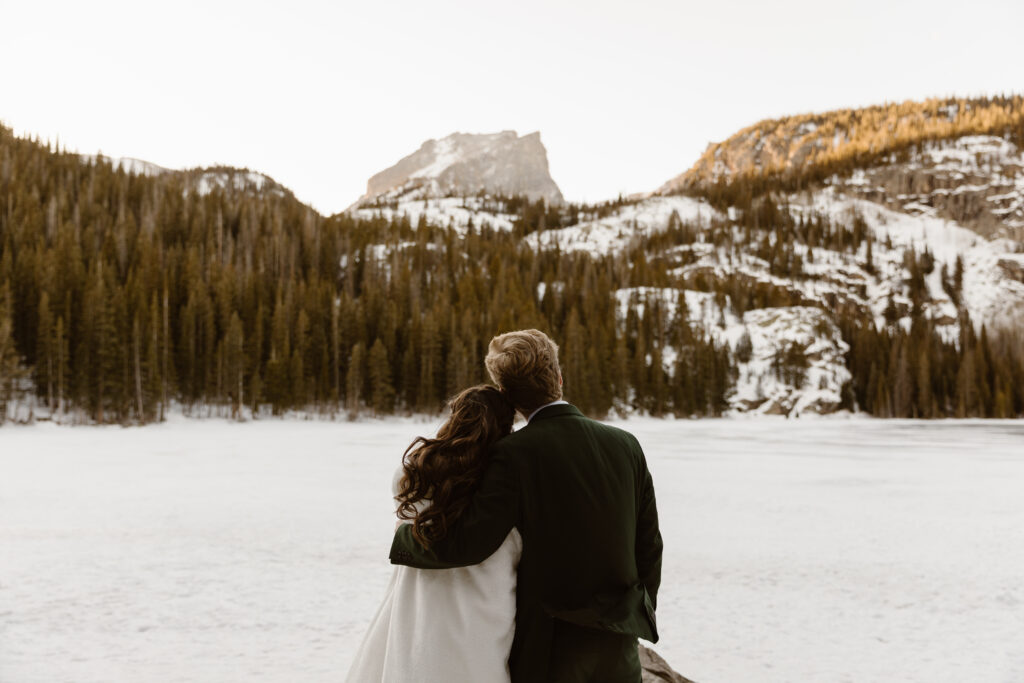 Bride and groom stand together embracing while looking out over a frozen lake and snowy mountains during a winter elopement in Rocky Mountain National Park.