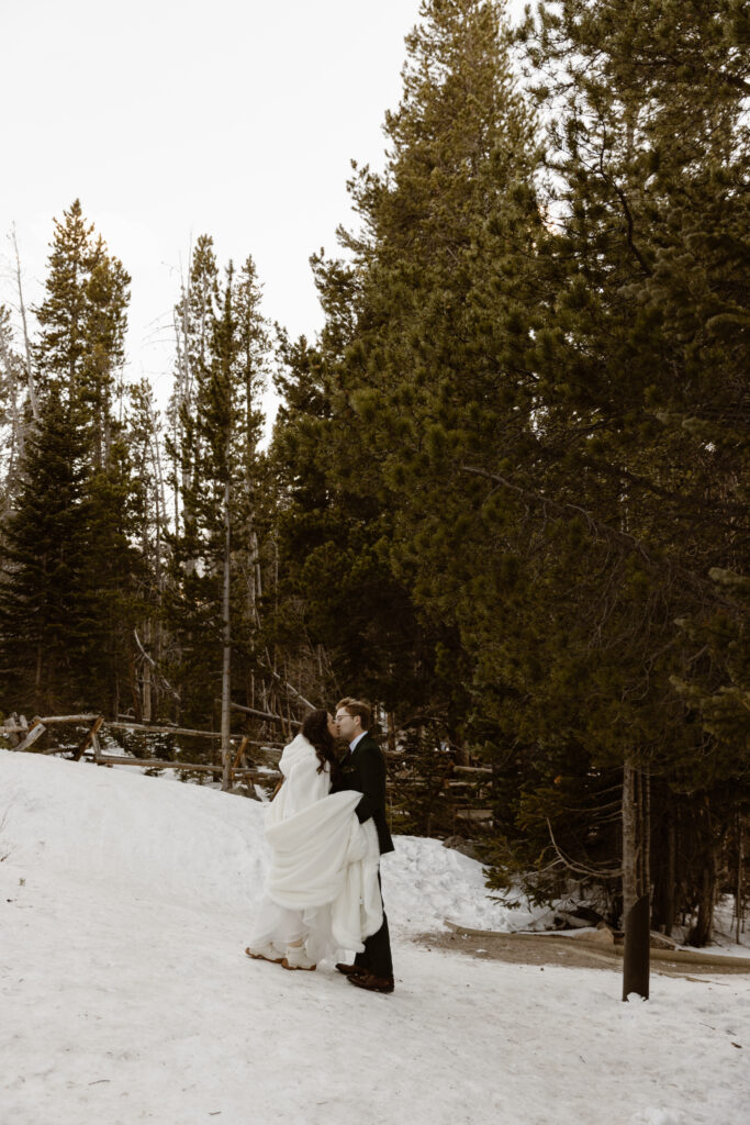 Bride and groom kiss on a snowy forest trail during a winter elopement in Rocky Mountain National Park surrounded by tall evergreen trees.