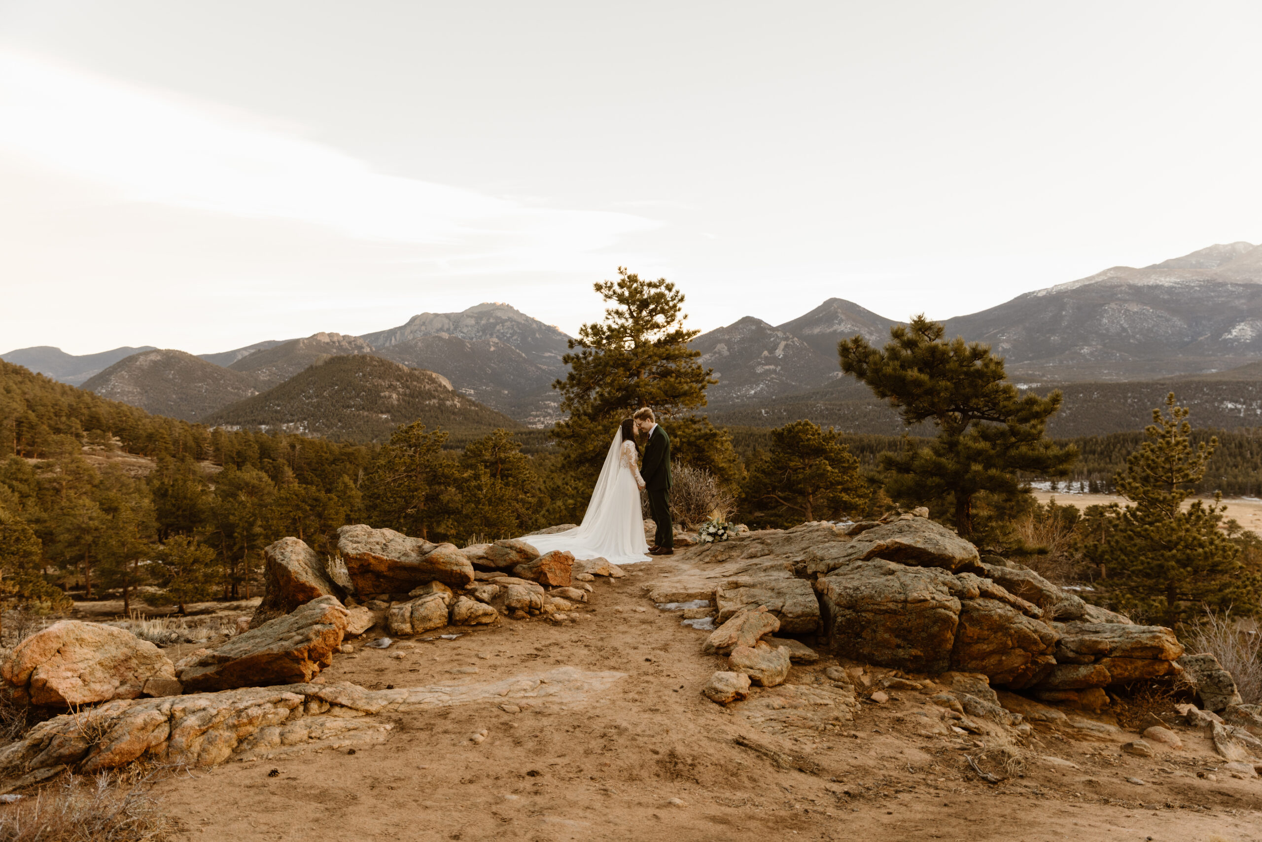 Couple stands together on a rocky overlook during a winter elopement in Rocky Mountain National Park, surrounded by mountains and pine trees.