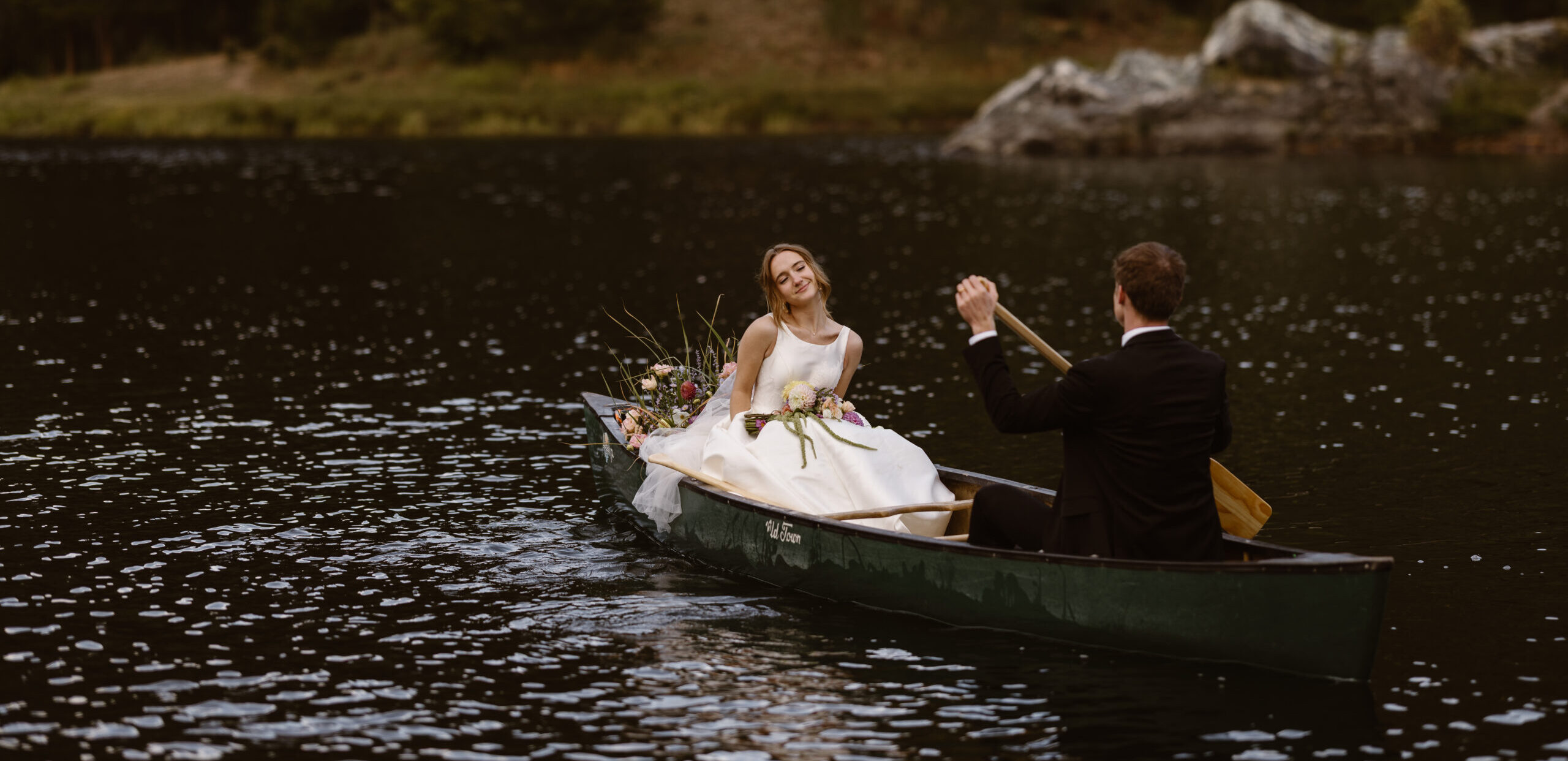Bride and groom sit together in a canoe on a calm lake, the groom paddling while the bride smiles softly surrounded by flowers in Dillon, Colorado.