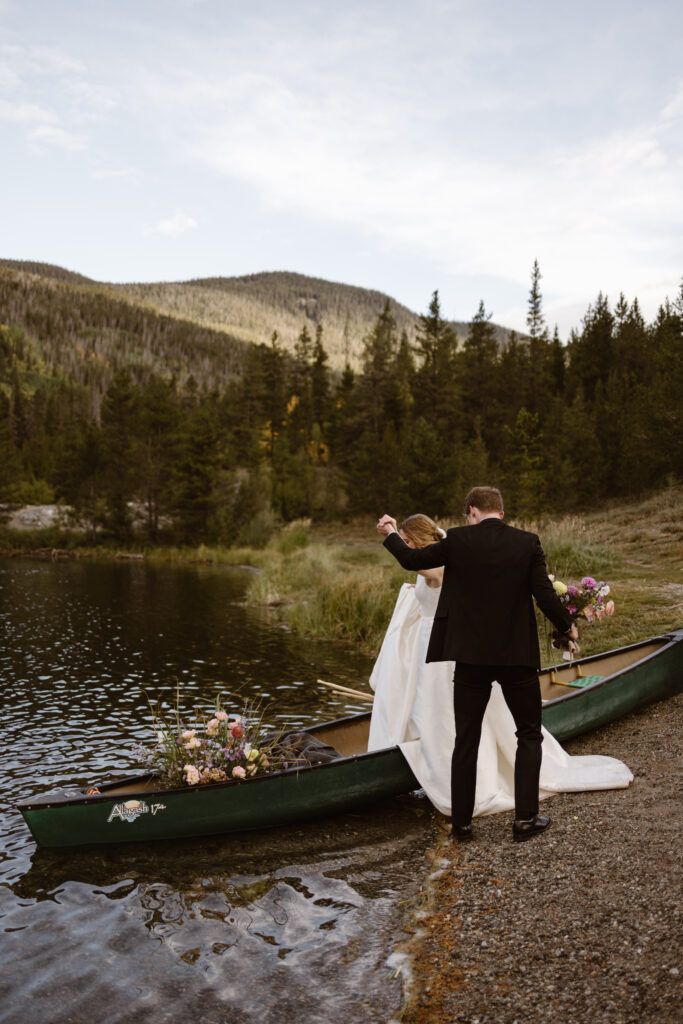 Couple steps into a green canoe along a mountain lake shoreline during an elopement in Dillon, Colorado, with forested hills in the background.