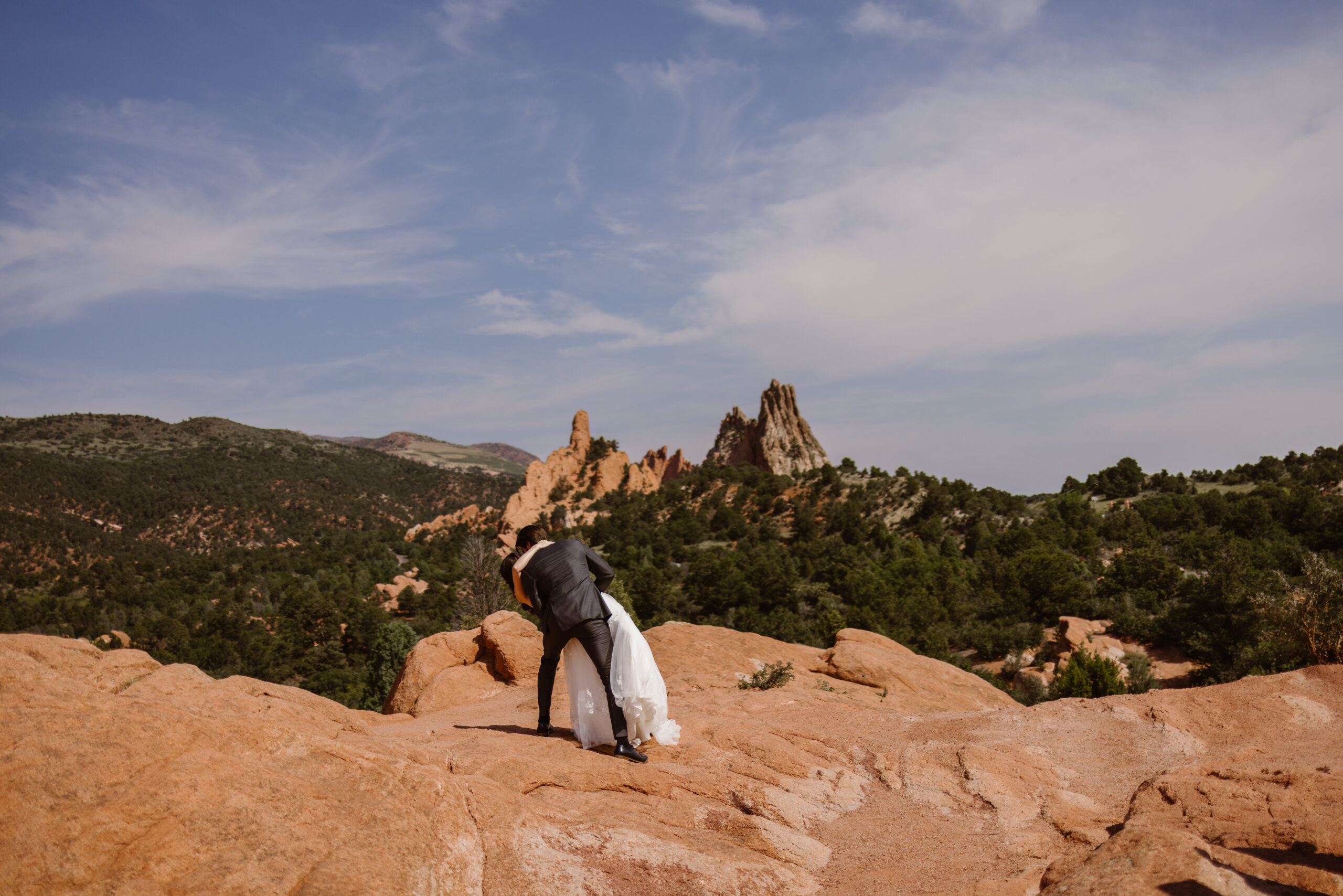 Bride and groom share their first kiss on the red rock formations at Garden of the Gods with sweeping Colorado mountain scenery and blue skies in the background.