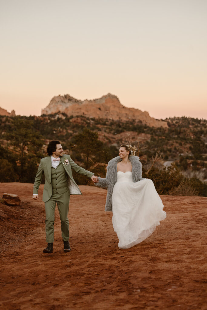 Bride and groom walk hand in hand across a red dirt trail during their Garden of the Gods elopement at sunset, smiling at each other with red rock formations in the background.