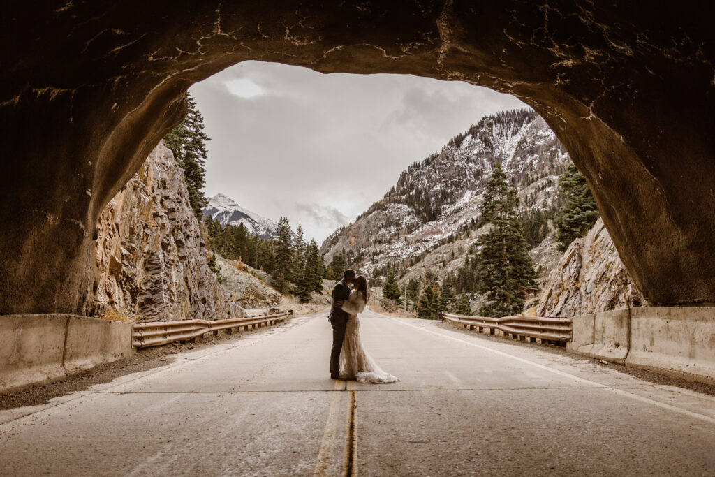 Bride and groom kiss in the middle of a mountain road framed by a rocky tunnel, with snow-dusted peaks and trees in the background in Ouray, Colorado.