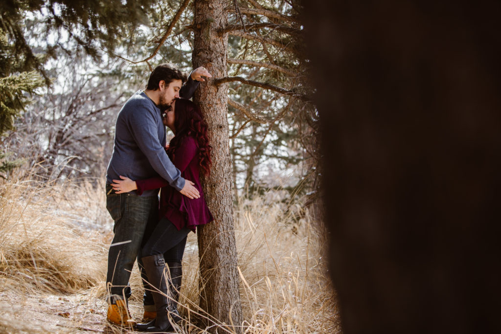 Man kissing his fiancé's forehead while leaning against a tree