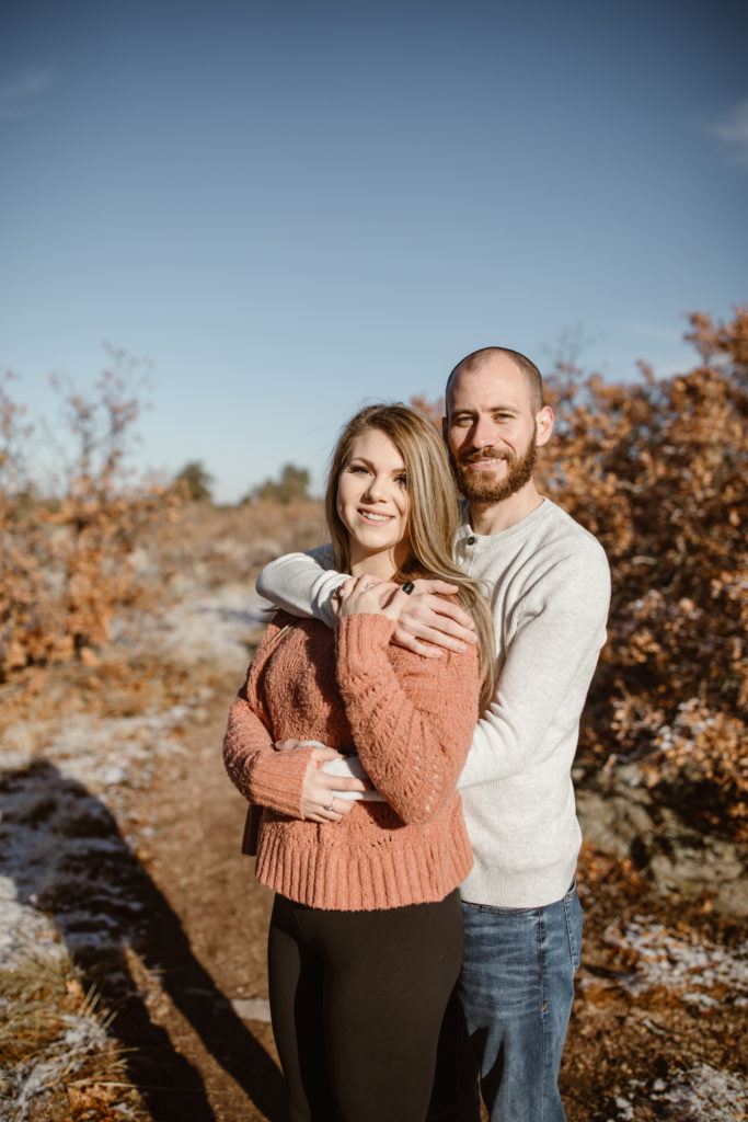 Man standing behind his fiancé both smiling and looking at the camera