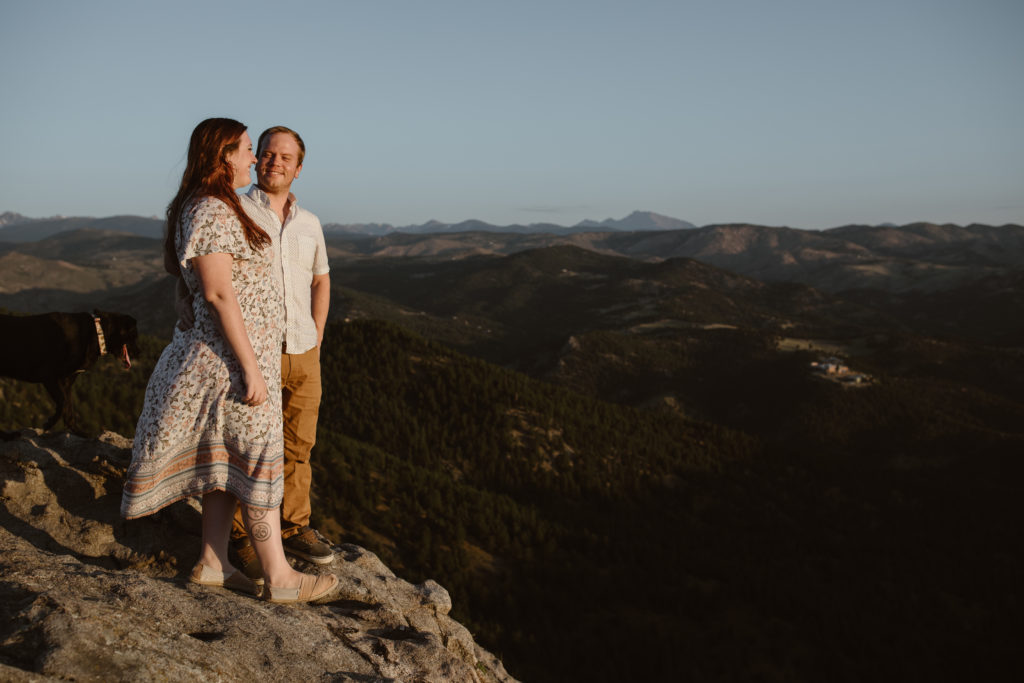 Couple with arms around each other on a rock in the mountains. Man is looking at his fiancé while she looks at the view