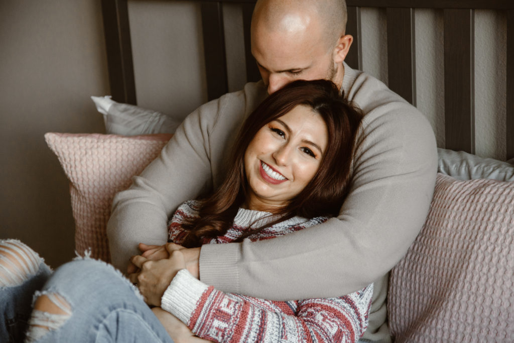 Couple cuddled on their bed, man's arms around fiancé who is smiling at the camera