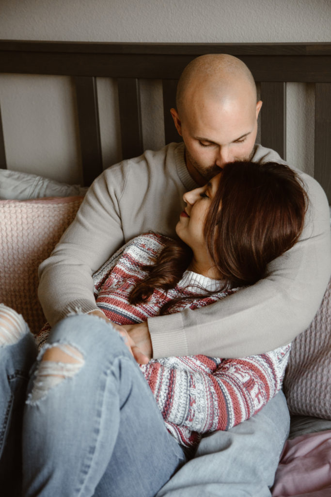 Couple cuddled on their bed, man giving his fiancé a kiss on the forehead
