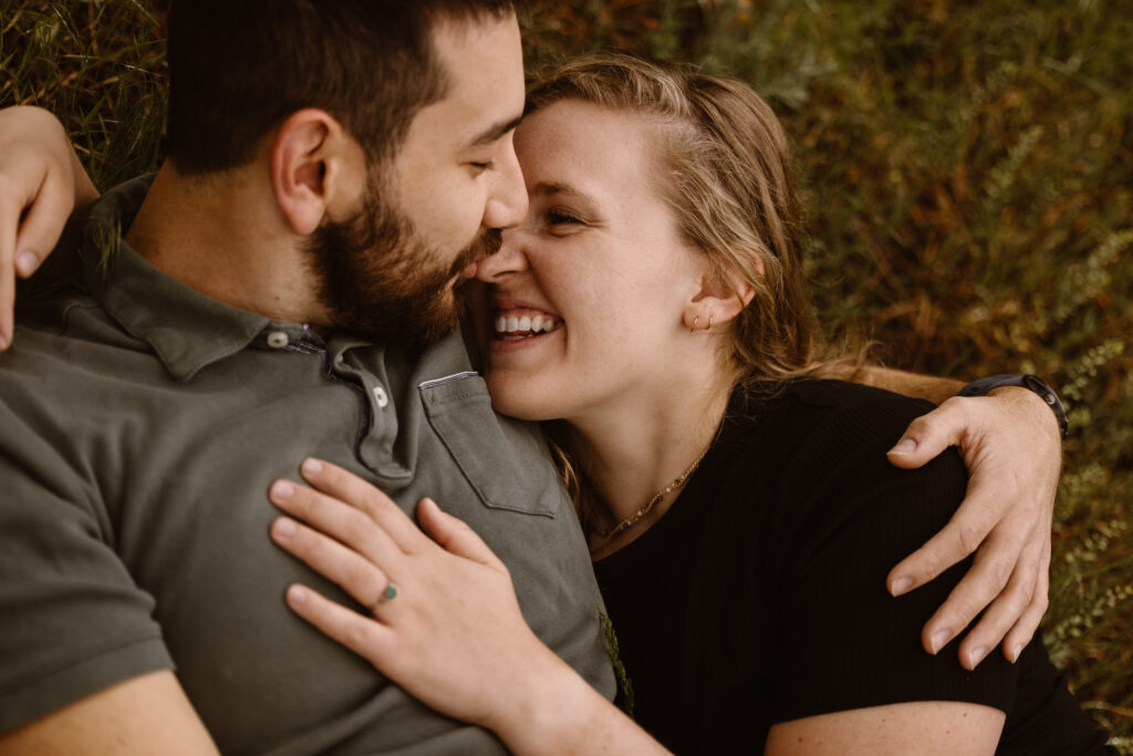 Couple laying on the ground cuddled up. Man is kissing his fiancé on the nose