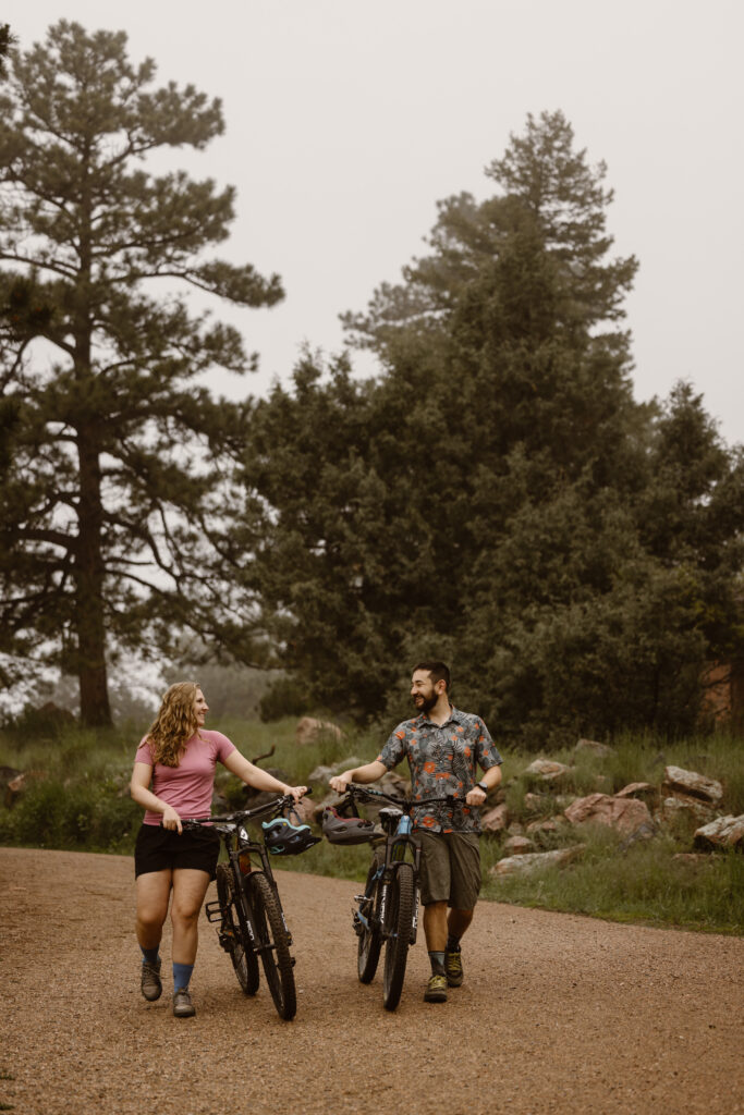 Engaged couple walking their mountain bikes smiling and looking at each other