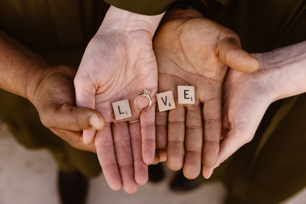 Couple holding Scrabble tiles spelling out "love" with the woman's wedding ring in place of the "o"
