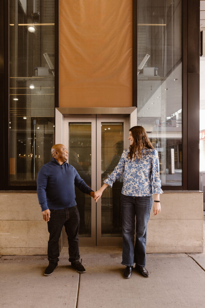 Couple standing in front of a glass elevator holding hands standing apart