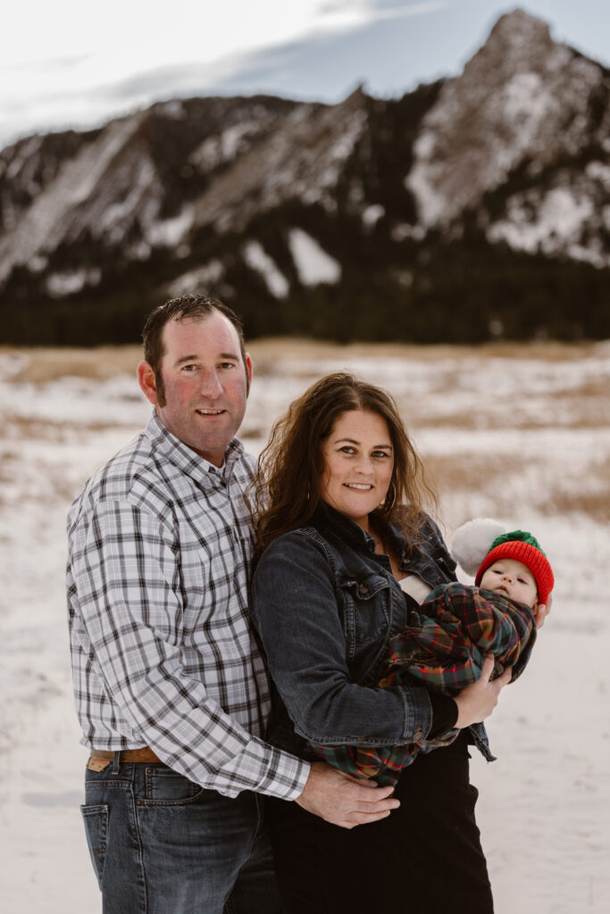 Husband and wife holding their newborn together with snowy Flatirons in the background