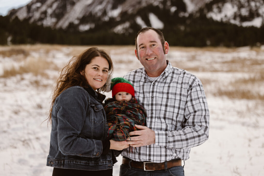 Husband and wife holding their newborn together with snowy Flatirons in the background