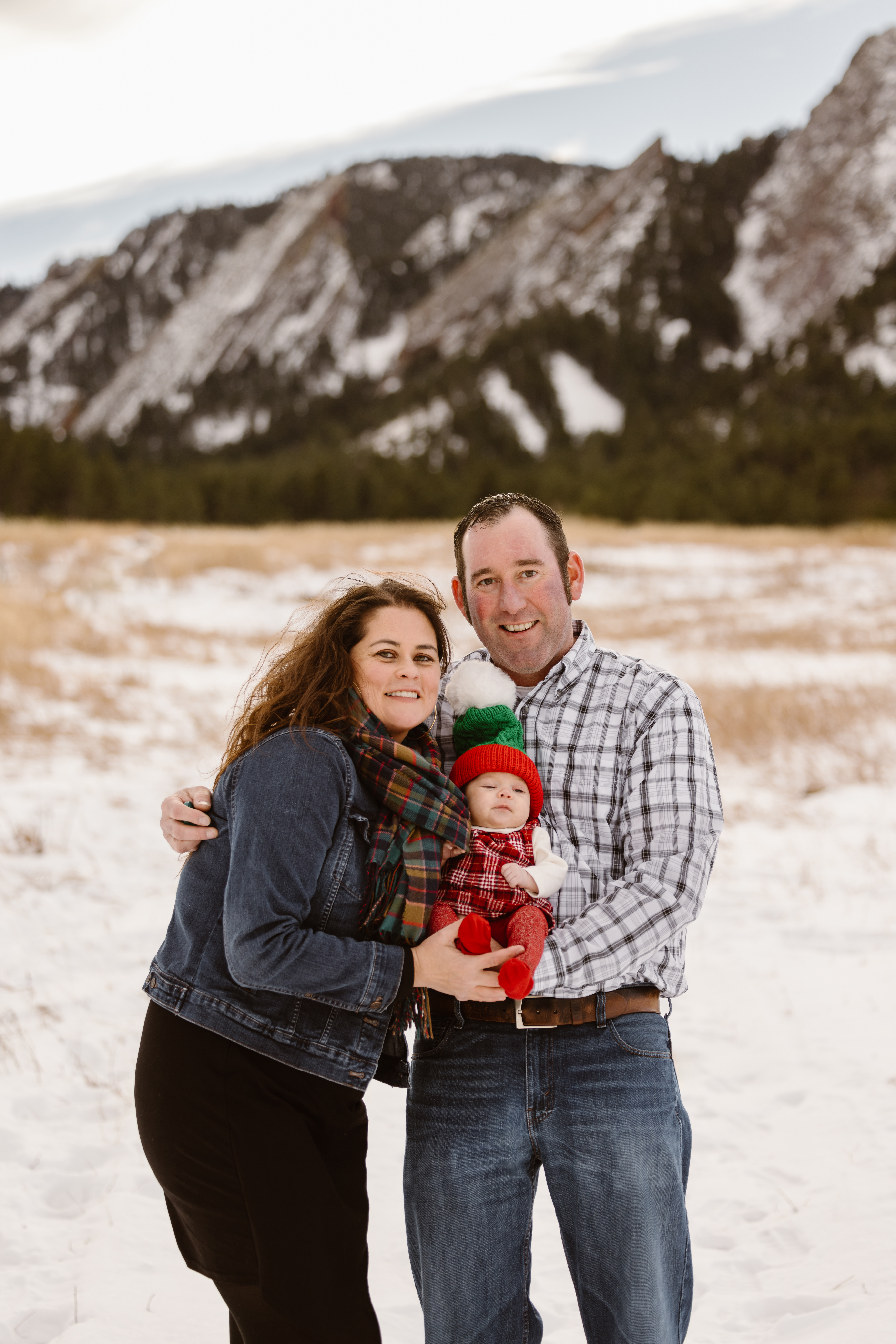 Husband and wife holding their newborn together with snowy Flatirons in the background