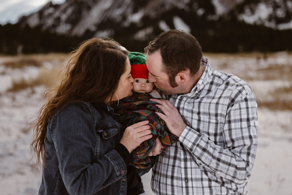 Husband and wife each kissing a cheek of their newborn daughter with snowy Flatirons in the background