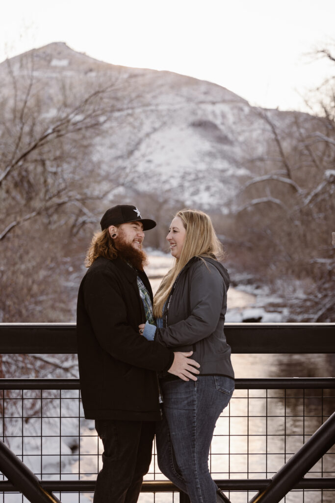 Couple cuddled together on a bridge over a river with a snowy river and mountain in the background