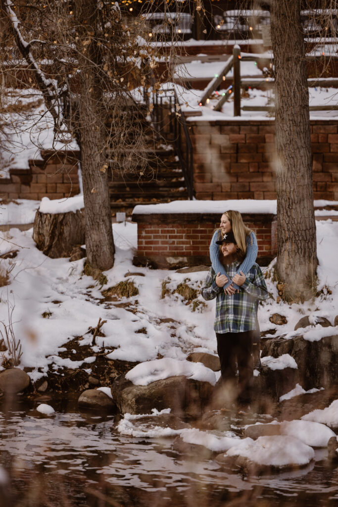 Couple cuddled together, woman behind her boyfriend arms wrapped around his shoulders next to a river with snow
