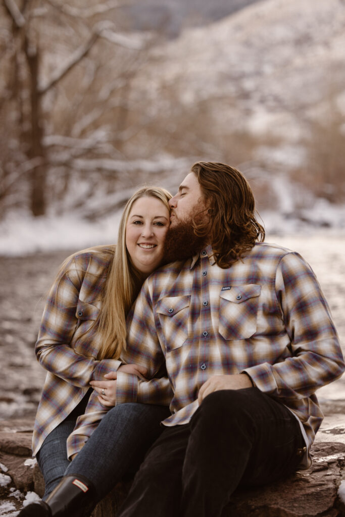 Couple sitting cuddled up next to each other, man kissing his girlfriend's temple, next to a river with snow