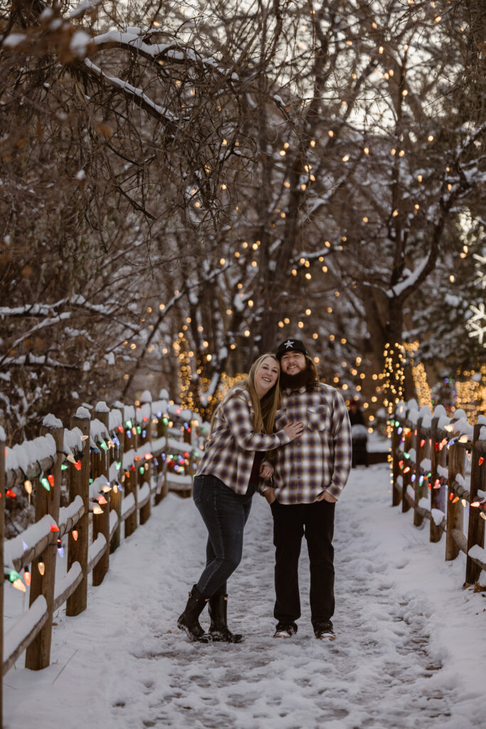 Couple on a snowy sidewalk with Christmas lights all over, woman looking at her boyfriend with her hand on his chest