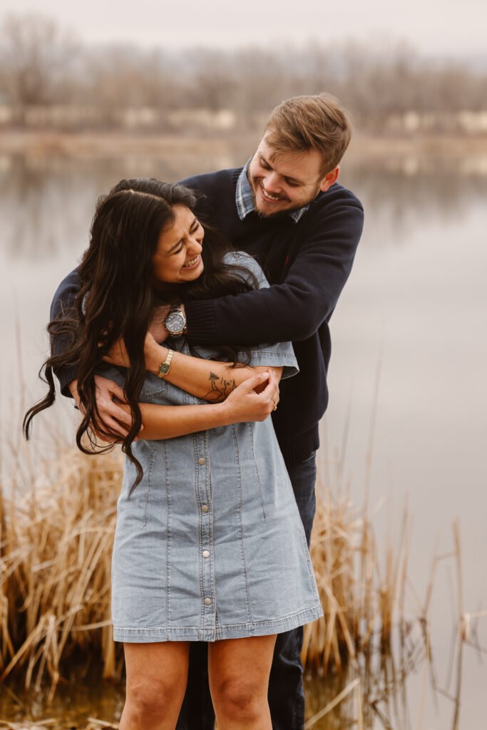 Man behind his fiancé with his arms wrapped around her, they are both laughing