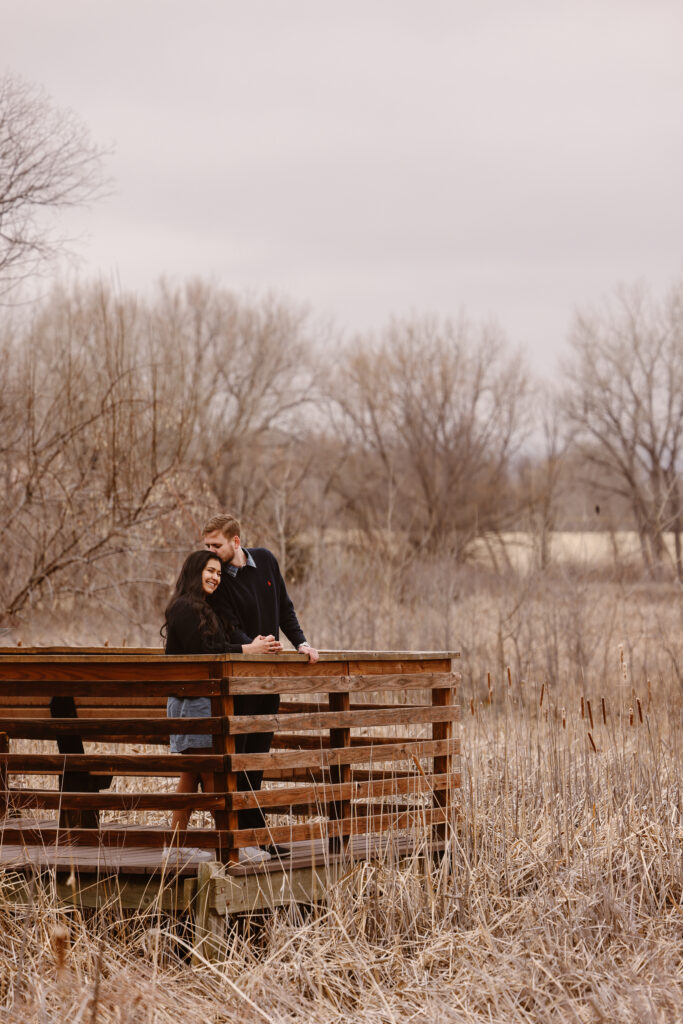 Man kissing his fiancé on the head while standing on a bridge on a lake