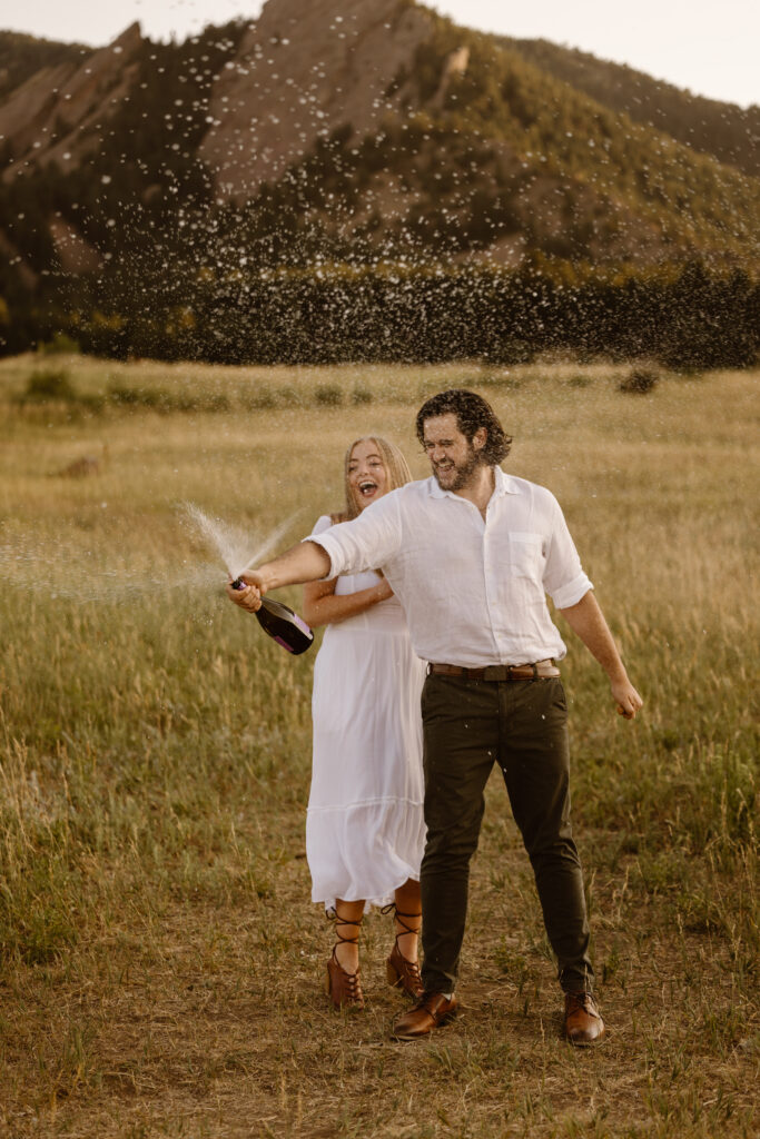 Playful engagement photo of a couple popping champagne with the Flatirons in Boulder, Colorado as the backdrop.