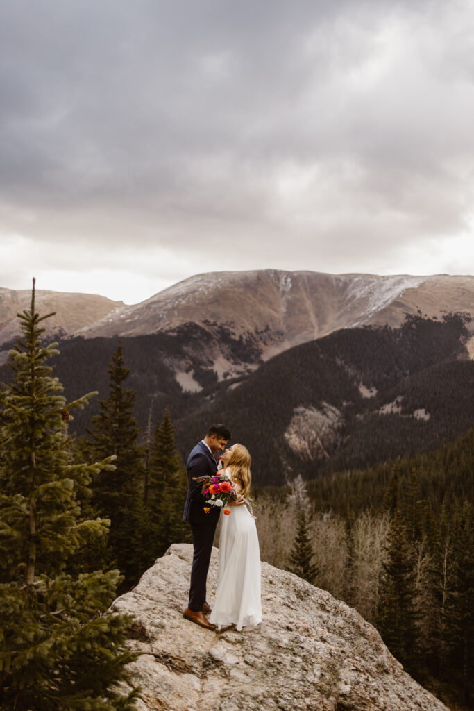 Bride and groom share a romantic kiss after exchanging vows during an intimate Winter Park elopement on a rocky mountain overlook surrounded by evergreen trees.