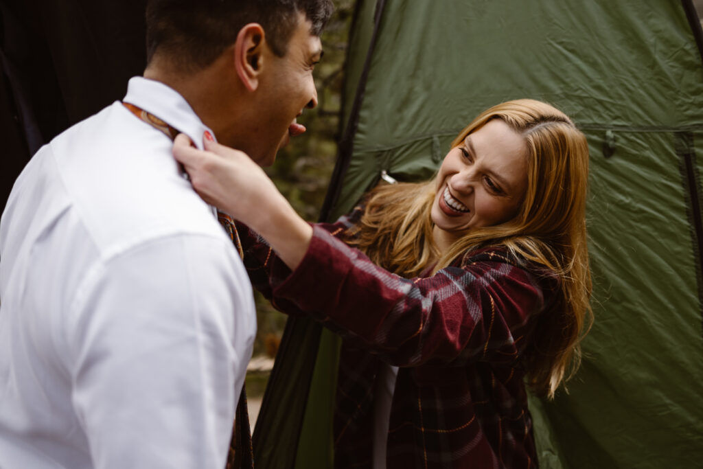 Partner laughing while helping the groom adjust his tie during a Winter Park elopement, with a green tent in the background.