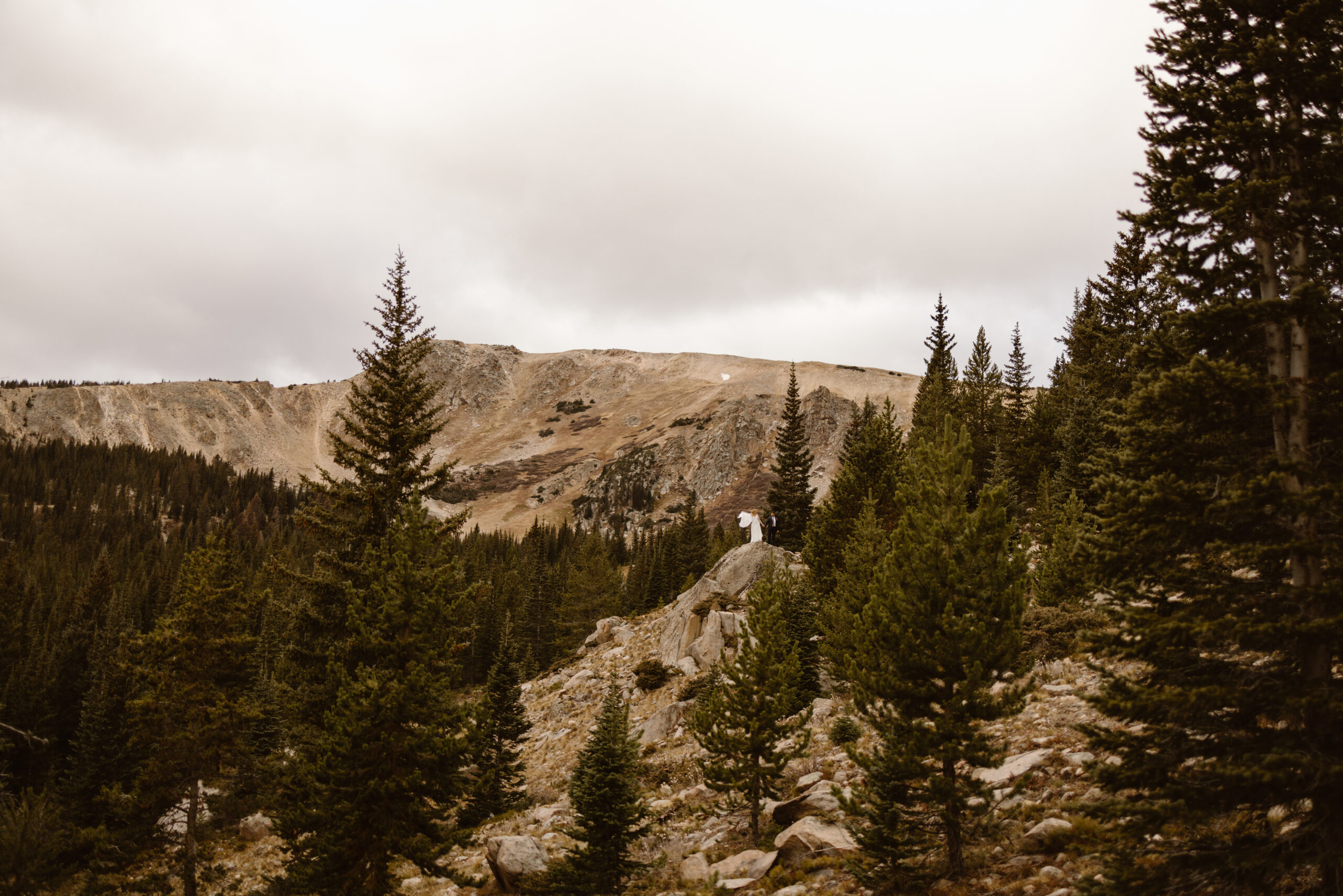 A newly married couple stands together on a rocky mountain overlook during their Winter Park, Colorado elopement, surrounded by evergreen trees and expansive alpine scenery under an overcast sky.