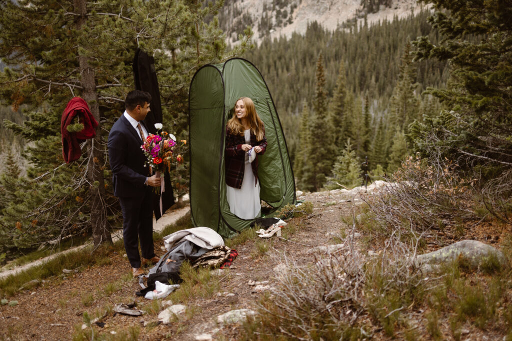 Bride smiles while stepping out of a green changing tent as her partner holds a colorful bouquet during a Winter Park elopement in the Colorado mountains.