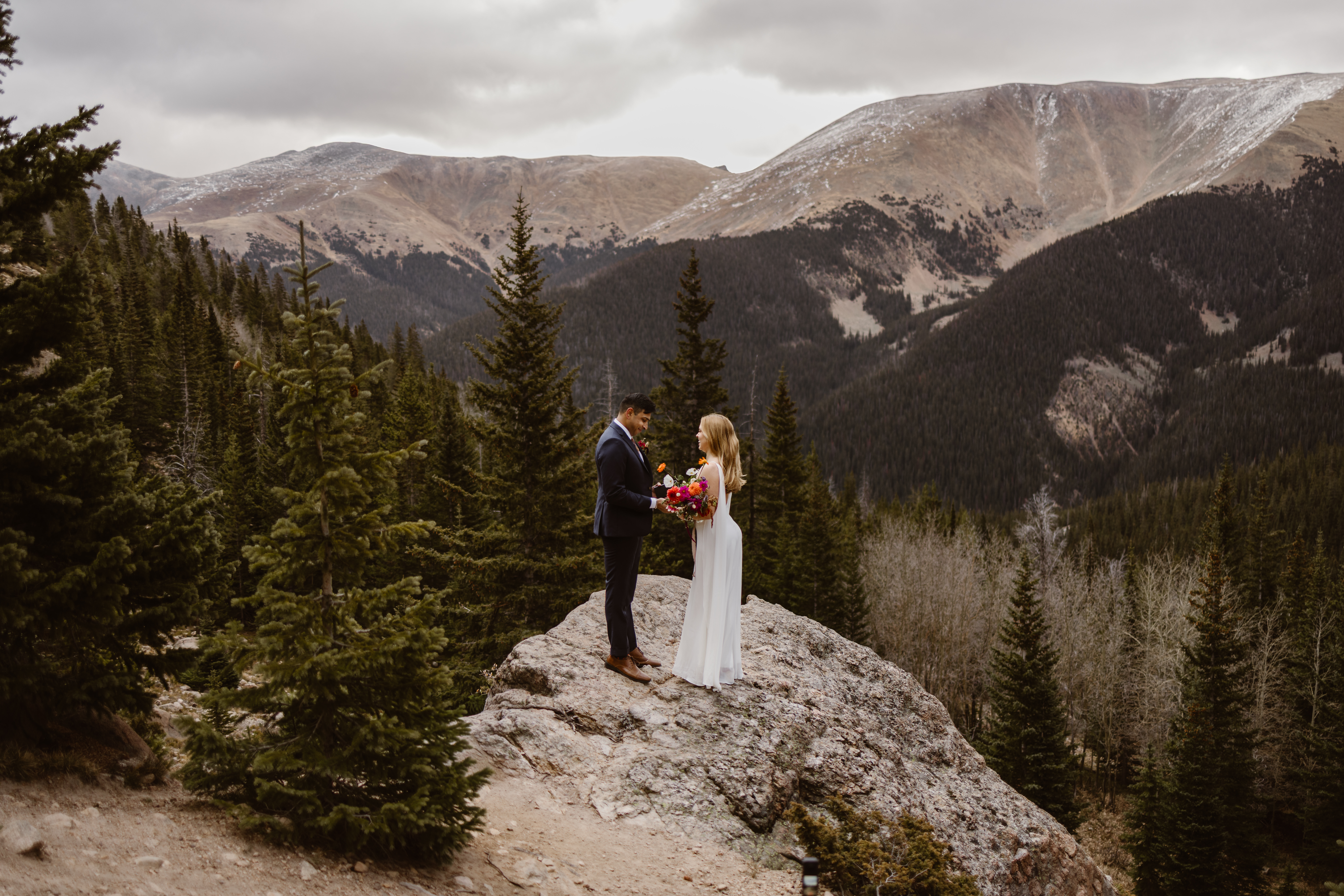 Groom reading his vows to his partner during a Winter Park elopement, standing together on a mountain overlook surrounded by evergreen trees and Colorado peaks.