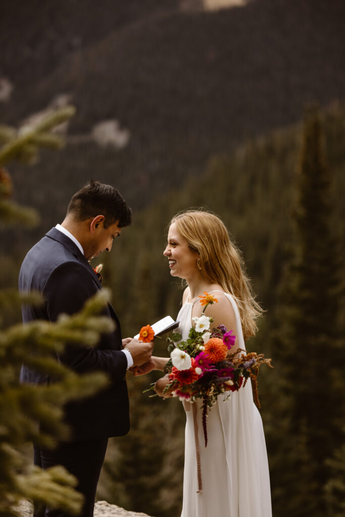 Bride smiles and laughs as her partner reads his vows during a Winter Park elopement in the Colorado mountains, holding a colorful wildflower bouquet.
