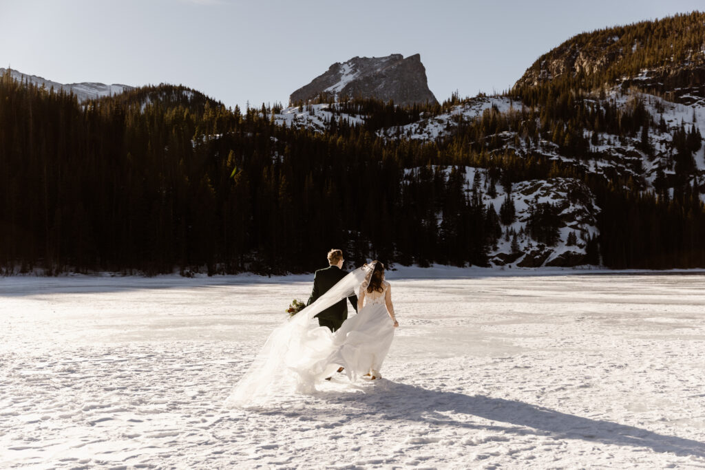 Bride and groom walking across a frozen lake in Rocky Mountain National Park during their Colorado winter elopement with snow-covered peaks in the background.