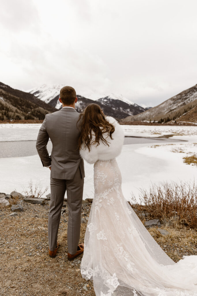 Bride and groom standing beside a partially frozen alpine lake during their Colorado winter elopement with snow-covered mountains in the background.