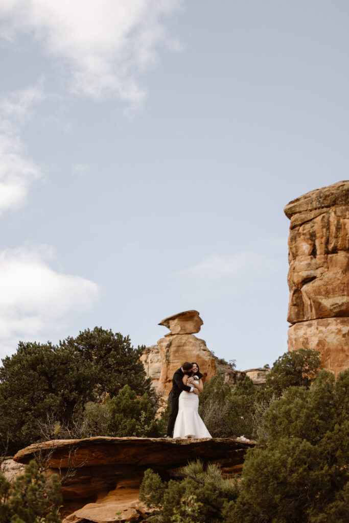Bride and groom embracing on a red rock ledge during their Colorado elopement with desert cliffs and blue sky in the background.