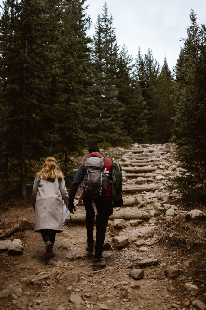Couple hiking up a rocky forest trail in Colorado while carrying gear on the way to their mountain elopement ceremony.